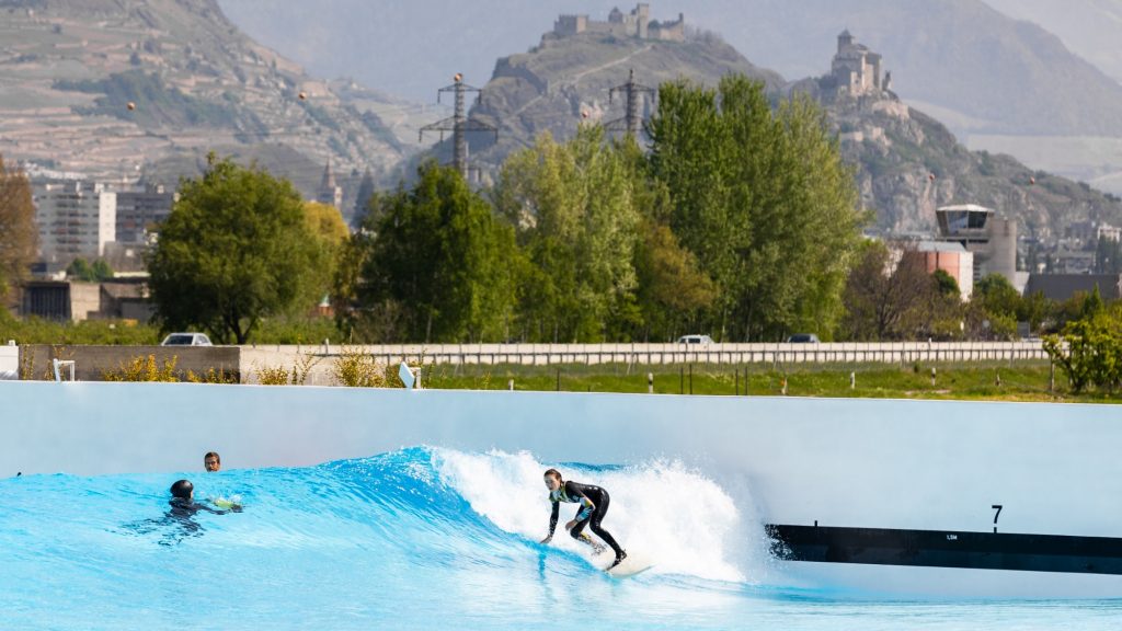 Travel to a wave pool destination such as switzerland is becoming more popular. Image shows surfer with alps as a backdrop